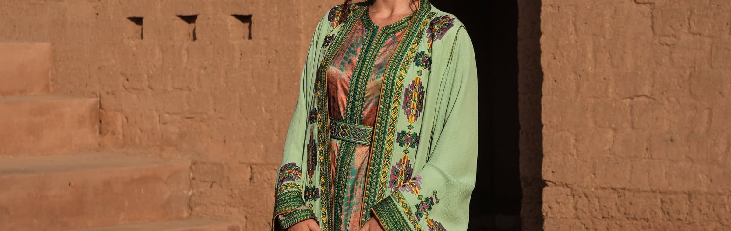 Anise green silk Moroccan kaftan with an embroidered placket against a Marrakech adobe wall